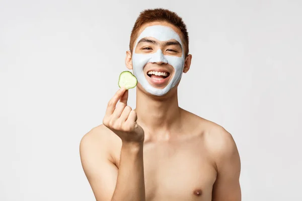 Bellezza, cura della pelle e concetto di spa. Ritratto di uomo asiatico allegro e sorridente con busto nudo, maschera viso applicata per rimuovere macchie, mostrando cetriolo, sfondo bianco in piedi — Foto Stock