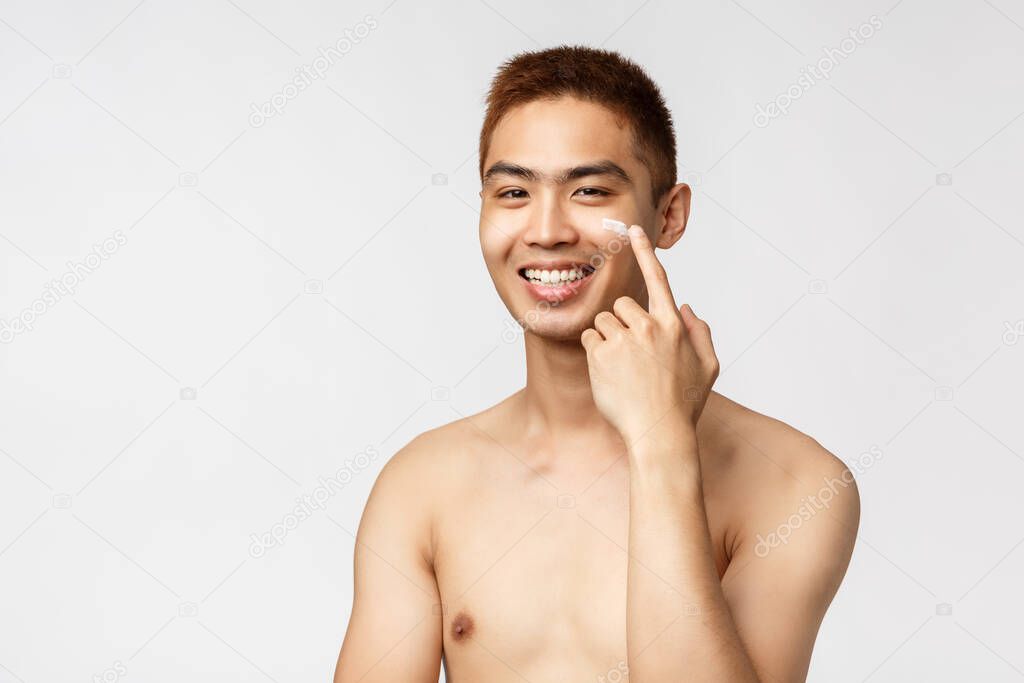 Beauty, people and leisure concept. Close-up portrait of pleased smiling asian man with naked torso, apply skincare routine cream on face and grinning delighted, white background