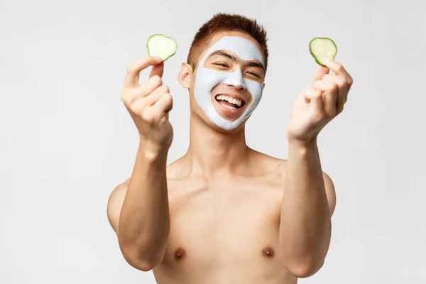 Skönhet, hudvård och spa-koncept. Porträtt av glad snygg asiatisk naken man som visar gurka bitar och leende, avkopplande hemma, tillämpa ansiktskräm, kosmetika mask, vit bakgrund — Stockfoto