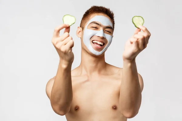 Bellezza, cura della pelle e concetto di spa. Ritratto di felice bell'uomo nudo asiatico che mostra pezzi di cetriolo e sorridente, rilassante a casa, applicare la crema per il viso, maschera cosmetica, sfondo bianco — Foto Stock