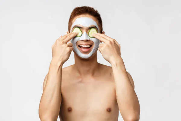 Ομορφιά, περιποίηση δέρματος και σπα. Πορτρέτο του ενθουσιώδη, χαλαρή ασιατικό γυμνό άνδρα απολαμβάνοντας τα Σαββατοκύριακα, εφαρμόστε ακμή θεραπεία προσώπου μάσκα και να τεθεί σε αγγούρια μάτια, χαμογελώντας ευρέως, λευκό φόντο — Φωτογραφία Αρχείου