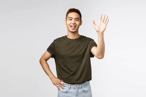 Mensen, emoties en levensstijl concept. Portret van vriendelijke vrolijke aziatische man die gedag zegt, welkomstpersoon als zwaaiende hand, informele begroeting, hallo leuk om je te ontmoeten, staande grijze achtergrond — Stockfoto