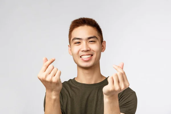 Люди, разные выражения и образ жизни. Симпатичный азиатский парень со стильной стрижкой, показывающий корейские пальчики сердца и улыбающийся милый, выражающий симпатию или вроде того, стоящий на сером фоне — стоковое фото