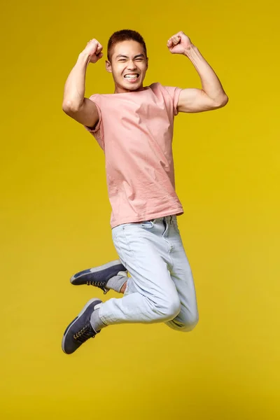 Verão férias, educação e estilo de vida conceito. Vertical tiro de feliz atlético asiático cara, flex bíceps e sorrindo otimista, pulando de alegria, triunfando, celebrando a vitória, fundo amarelo — Fotografia de Stock