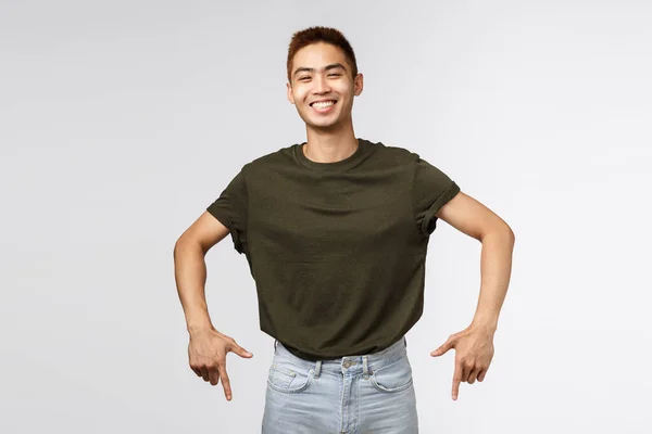 Portret van een vrolijke Aziatische man in een grijs t-shirt, vrolijk lachend en glimlachend, wijzend met de vingers naar beneden, uitnodigend om te klikken link, abonneren of uit te vinden aanbieding details op de onderste banner, grijze achtergrond — Stockfoto