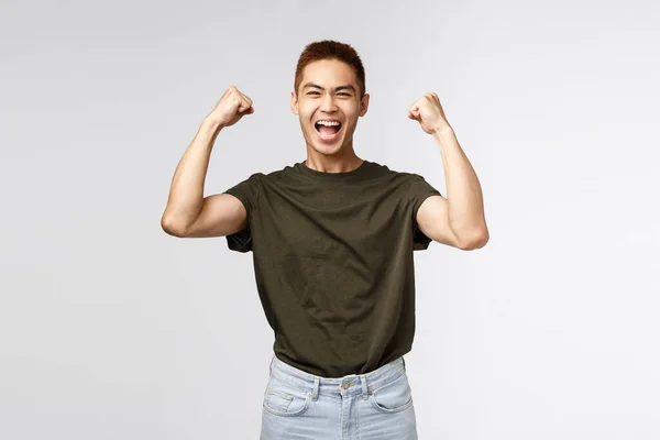 Concurso, premio y concepto de personas. Retrato de feliz, triunfante joven asiático levantando las manos, bomba de puño celebrando la victoria, lograr el éxito y ganar, sonriendo optimista, fondo gris — Foto de Stock