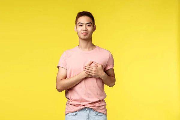 Lidé, různé emoce a životní styl. Usmívající se dotkl asijský muž se stylovým účesem, růžové tričko, držet se za ruce na srdci a vypadající fotoaparát potěšen, jít na dovolenou, žluté pozadí — Stock fotografie