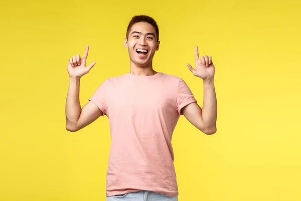 Retrato de joven chico asiático alegre en camiseta rosa, señalando los dedos hacia arriba, mostrando el camino al producto, sonriendo divertido y feliz, dar consejos, recomendar artículo, fondo amarillo — Foto de Stock