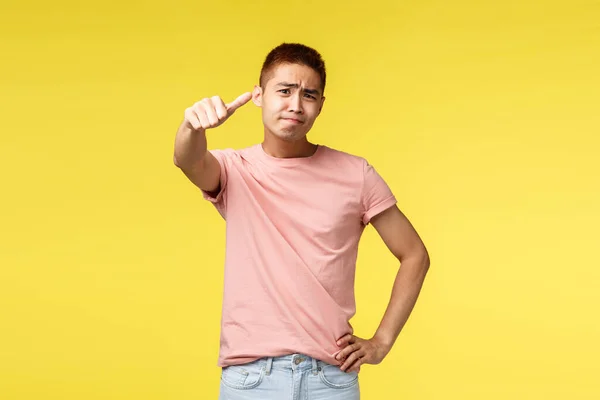Retrato de cético desagradado asiático homem em rosa t-shirt, julgar, expressar própria opinião para coisa ruim, taxa de produto é médio, polegar para baixo e sorridente decepcionado, fundo amarelo — Fotografia de Stock
