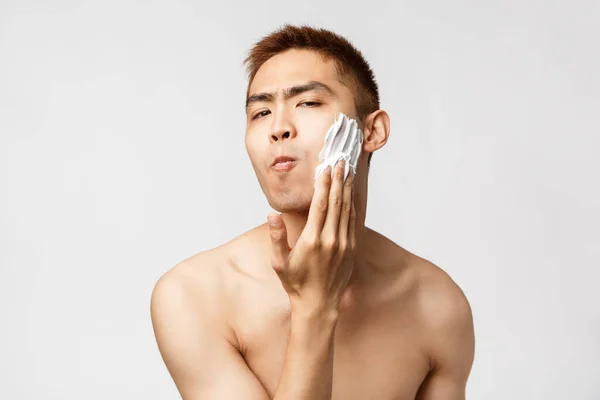 Beauty, people and hygiene concept. Portrait of funny handsome young man with naked torso, looking at bathroom mirror, apply shaving cream to face, standing white background — Stock Photo, Image