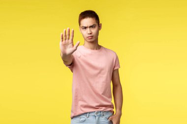 Judgemental and skeptical young asian guy pull one hand in prohibition, tell no, stop it, serious looking camera, frowning and grimacing bothered, annoyed, express disagreement, yellow background clipart