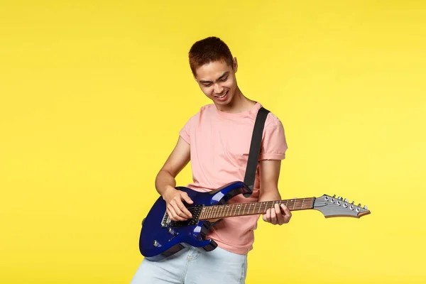 Lifestyle, leisure and youth concept. Portrait of happy, cool and stylish asian guy playing in band, enjoying perfoming on electric guitar, smiling enthusiastic, standing yellow background — Stock Photo, Image