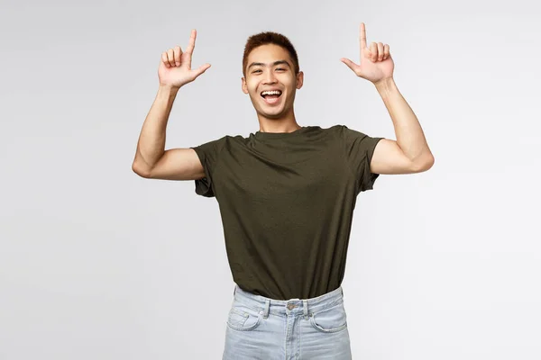 Portret van optimistische, blije Aziatische man in groen t-shirt die mensen uitnodigt om advertenties te bekijken, te lachen en te glimlachen opgewonden, vingers omhoog te wijzen, weg te wijzen naar banner, grijze achtergrond — Stockfoto