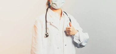 Covid19, koronavirüs, sağlık ve doktor konsepti. Genç kararlı Asyalı kadın doktor en kaliteli hastane ekipmanlarını garanti ediyor, baş parmağını kaldırıyor, beyaz önlük ve stetoskop giyiyor, tıbbi maske takıyor.