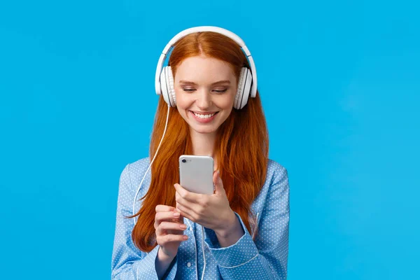 Technologiesucht, Lebensstil und Frauenkonzept. fröhlich gut aussehende rothaarige Frau in Nachthemd, mit großen Kopfhörern Musik hören, lächelnd Lieder in Smartphone-App auswählen — Stockfoto
