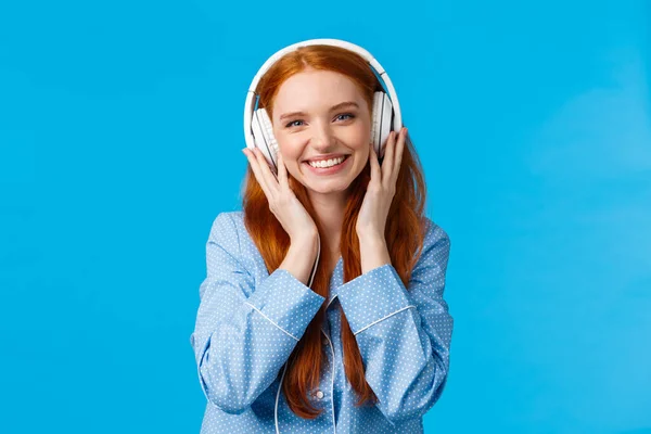 Cintura-para arriba tiro alegre bastante foxy chica caucásica escuchar música en auriculares grandes, sonriendo complacido, amigos de salida solo, usando ropa de dormir, de pie fondo azul encantado — Foto de Stock