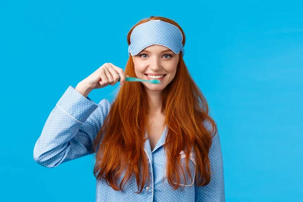 Habits, hygiene and lifestyle concept. Cute feminine redhead european woman in nightwear, sleep mask smiling and brushing teeth with toothbrush, standing blue background — 스톡 사진