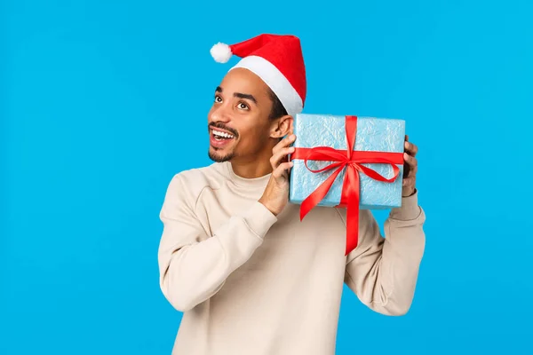 Hmm interesante lo que hay dentro. Curioso y entusiasta chico afro-americano feliz en sombrero de navidad santa, sacudiendo caja de regalo cerca de la oreja para escuchar y adivinar qué es, sonriendo, celebrando las vacaciones de invierno — Foto de Stock