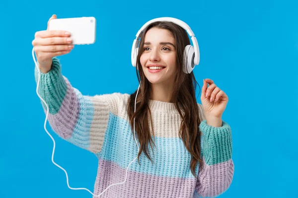 Retrato en la cintura linda mujer joven bonita con el pelo largo y oscuro, sonrisa alegre, usar auriculares, tomar selfie en el teléfono inteligente, levantar la mano con el teléfono y posar, de pie fondo azul — Foto de Stock