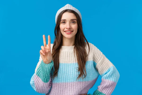 Скажи сыр. Симпатичная брюнетка-кавказка в зимней шляпе и свитере, наслаждающаяся рождественскими праздниками, отдыхом на горнолыжном курорте, показывающая знак мира и улыбающуюся камеру счастливый, синий фон — стоковое фото