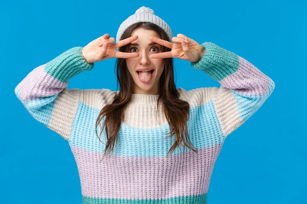 Будь собой. Привлекательная и игривая веселая, красивая кавказская девушка в зимней шляпе, свитере, показывающая язык и делающая диско, знаки мира над глазами, развлекающаяся на рождественских праздниках, голубой фон — стоковое фото