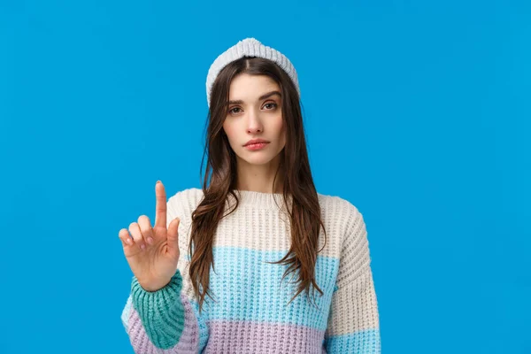 Regla número uno. Mujer asertiva de aspecto serio en suéter de invierno, sombrero, levantando un dedo en desaprobación, restricción o advertencia, prohibir hacer algo, gesto no tan rápido, fondo azul —  Fotos de Stock