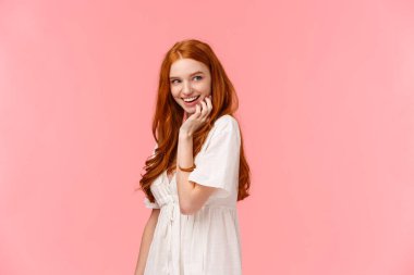 Waist-up portrait seducative and sensual, coquettish redhead flirty woman in white dress, glancing right smiling curiously, standing pink background intrigued, checking out something interesting