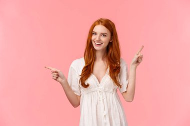 Happy alluring redhead female in white dress, helping out with fashion advices, pointing fingers left and right company banners, advertisements, smiling friendly and confident, showing what pick