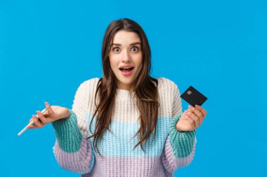 Şaşırdım ve merak ettim, kışın süveter giyen sevimli beyaz kadın, hayret içinde, kredi kartı ve akıllı telefon tutuyor, nakde çevrilmiş, mavi arka planda neşeli duruyor.
