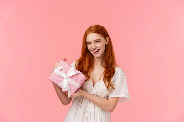 Emocionada descarada mujer pelirroja guapa en vestido blanco con estilo, sosteniendo regalo envuelto, preguntando qué hay dentro, sonriente descarado, dando regalo romántico para el día de San Valentín, fondo rosa —  Fotos de Stock