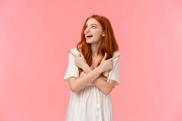 Dreamy attractive redhead female, looking around wondered and amused with happy smile, glance left as pointing sideways, left and right products, making final choice what buy, pink background — 스톡 사진