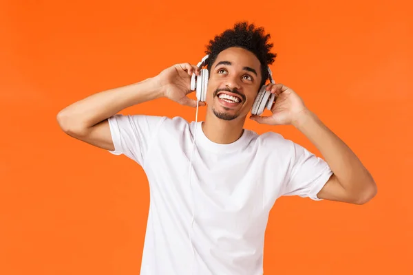 アフリカ系アメリカ人の男性が音楽を聴いたり、ヘッドフォンを手にしたり、笑顔で見上げたり、素晴らしいビートイヤフォンの楽しさを感じたり、オレンジの背景 — ストック写真