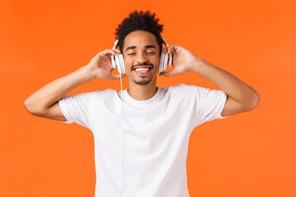 テクノロジー、ガジェットの概念。幸せな現代アフリカ系アメリカ人男性で白いtシャツに置くヘッドフォン、近い目と笑顔恍惚、お気に入りの音楽、良い音質、オレンジの背景を聞く — ストック写真