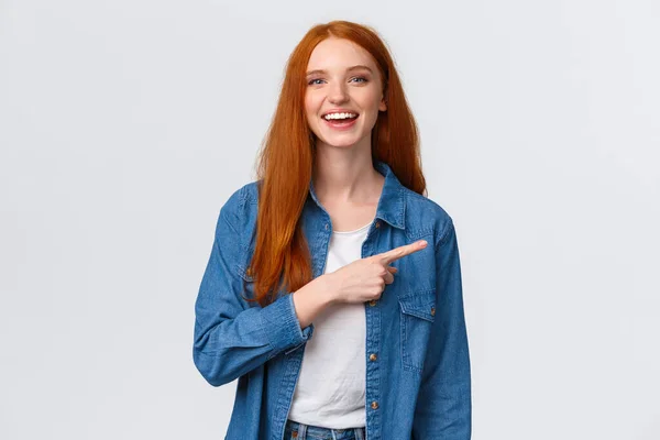 Despreocupada, amigable y feliz pelirroja extrovertida con camisa vaquera, señalando con el dedo hacia la derecha y riéndose de la cámara con sonrisa radiante, de pie fondo blanco, discutiendo, comunicando equipo — Foto de Stock