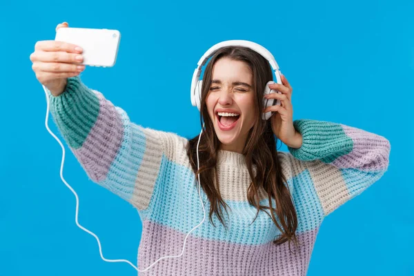 Joyeux, charismatique et souriant jeune femme magnifique en pull d'hiver, jouissant d'une musique impressionnante, prenant selfie comme écouter la chanson préférée dans les écouteurs, tenant smartphone, fond bleu — Photo