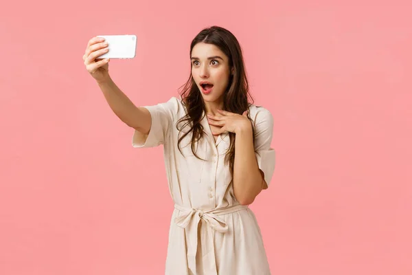 Girl making fascinated and surprised expression as talking to girlfriend on phone video-call. Charming brunette female in dress, taking selfie, gasping open mouth amused, standing pink background — Stock Photo, Image