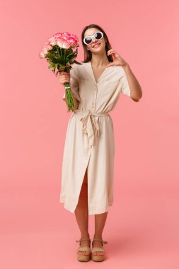 Tam boy dikey portre romantik ve şehvetli genç bir kadın elinde buket çiçekleri tutarak kalp işareti yapıyor. Sevgisini ifade ediyor ve güllerle şükran duyuyor.