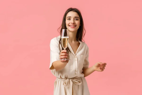 Here, make a toast. Cheerful romantic and alluring young girlfriend extending hand with glass, giving champagne to boyfriend, smiling joyfully, enjoying awesome party, pink background