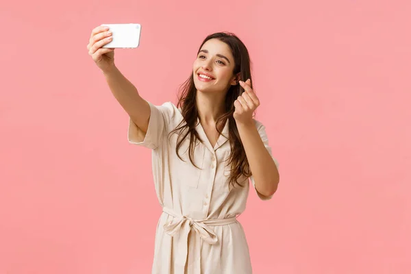 Веселая прекрасная молодая европейская женщина, использующая фильтр, чтобы сделать милое селфи, фотографируя на мобильную камеру, улыбаясь и показывая корейский жест сердца на смартфон, стоя розовый фон — стоковое фото
