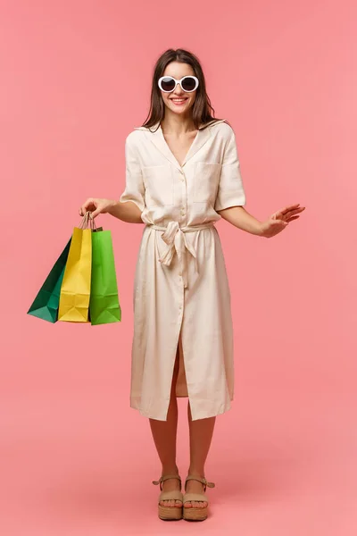 Retrato vertical de cuerpo entero elegante mujer guapa de vacaciones, de compras y divirtiéndose en centros comerciales, con gafas de sol, vestido ligero, llevar bolsas de la tienda, de pie fondo rosa sonriendo —  Fotos de Stock