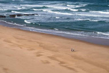 Monte Clerigo plajı boyunca yürüyen insanların siluetleri - Praia de Monte Clerigo - rüzgarlı ve güneşli bir günde Portekiz 'de dalgalar akarken