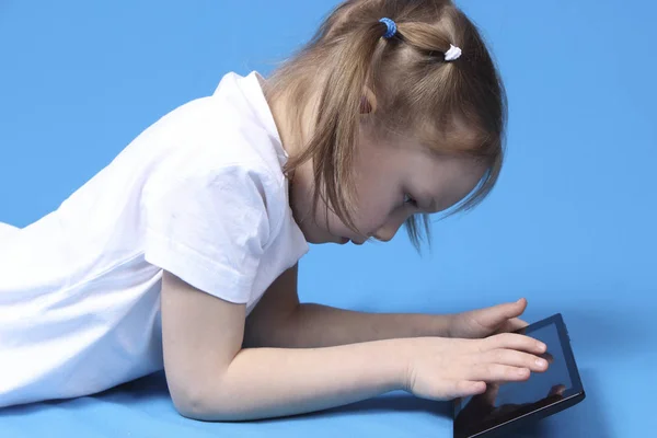 Flickan är liggande och spelar på tabletten. isolerad på blå bakgrund — Stockfoto