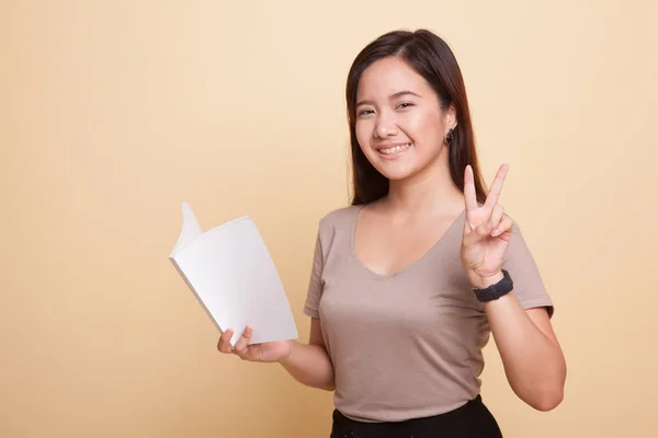 Junge asiatische Frau zeigt Siegerzeichen mit einem Buch. — Stockfoto