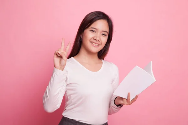 Junge asiatische Frau zeigt Siegerzeichen mit einem Buch. — Stockfoto