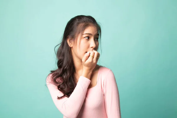 Unsure hesitant nervous young asian  woman biting her fingernails on cyan background