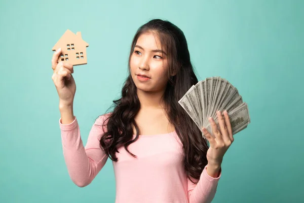 Happy young asian woman with house model and bunch of money banknotes on cyan background