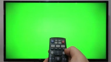 TV kumandasını gösteren ve yeşil ekranlı bir televizyonun önündeki düğmelere basan kişi