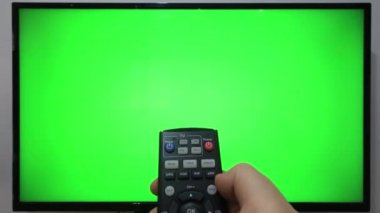 TV kumandasını gösteren ve yeşil ekranlı bir televizyonun önündeki düğmelere basan kişi