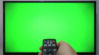 TV kumandasını gösteren ve yeşil ekranlı bir televizyonun önündeki düğmelere basan kişi
