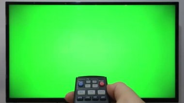 TV kumandasını gösteren ve yeşil ekranlı bir televizyonun önündeki düğmelere basan kişi